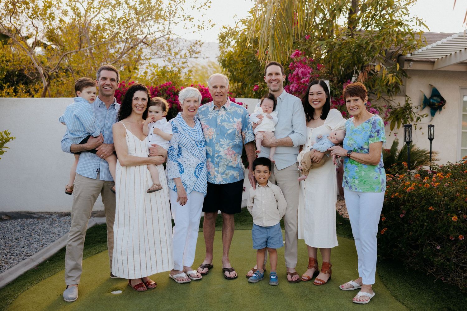 Multi-generational family posing together outdoors in a sunny backyard, smiling for a group portrait photo.