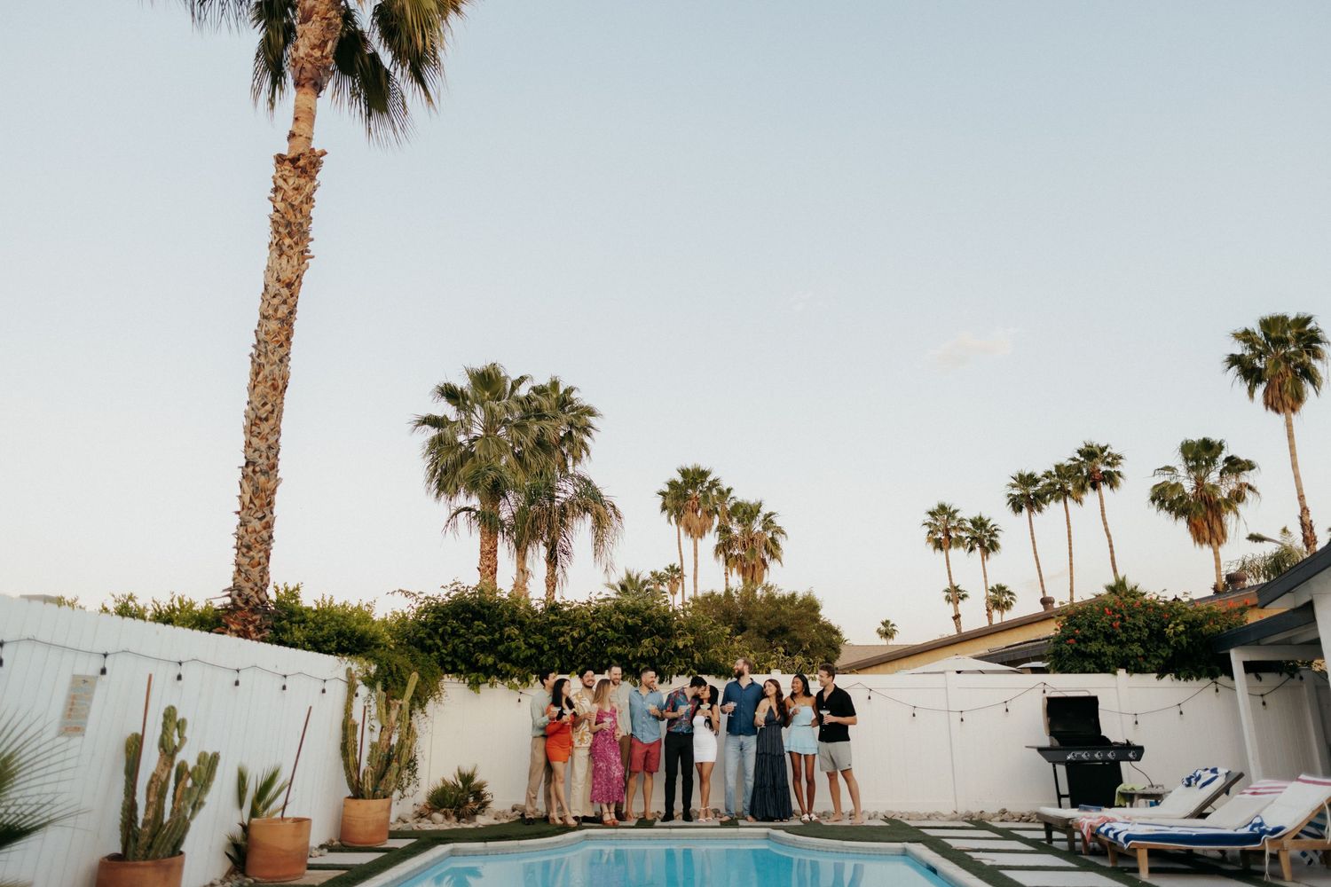 Group of friends posing by a pool surrounded by palm trees at a sunny backyard gathering in Palm Springs.