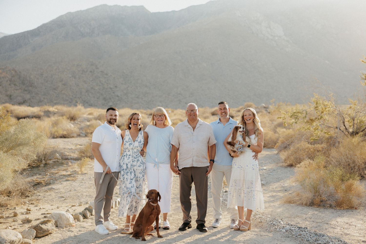 Family of six poses with a dog in a desert landscape with mountains in the background on a sunny day.