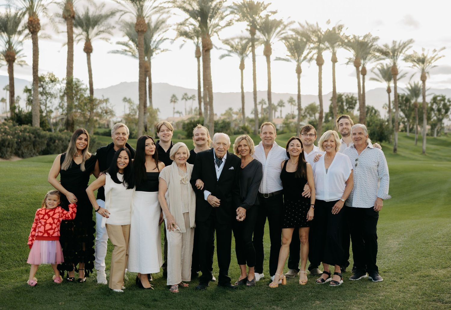 Large family group photo on a lush golf course with tall palm trees, posing together outdoors at dusk.