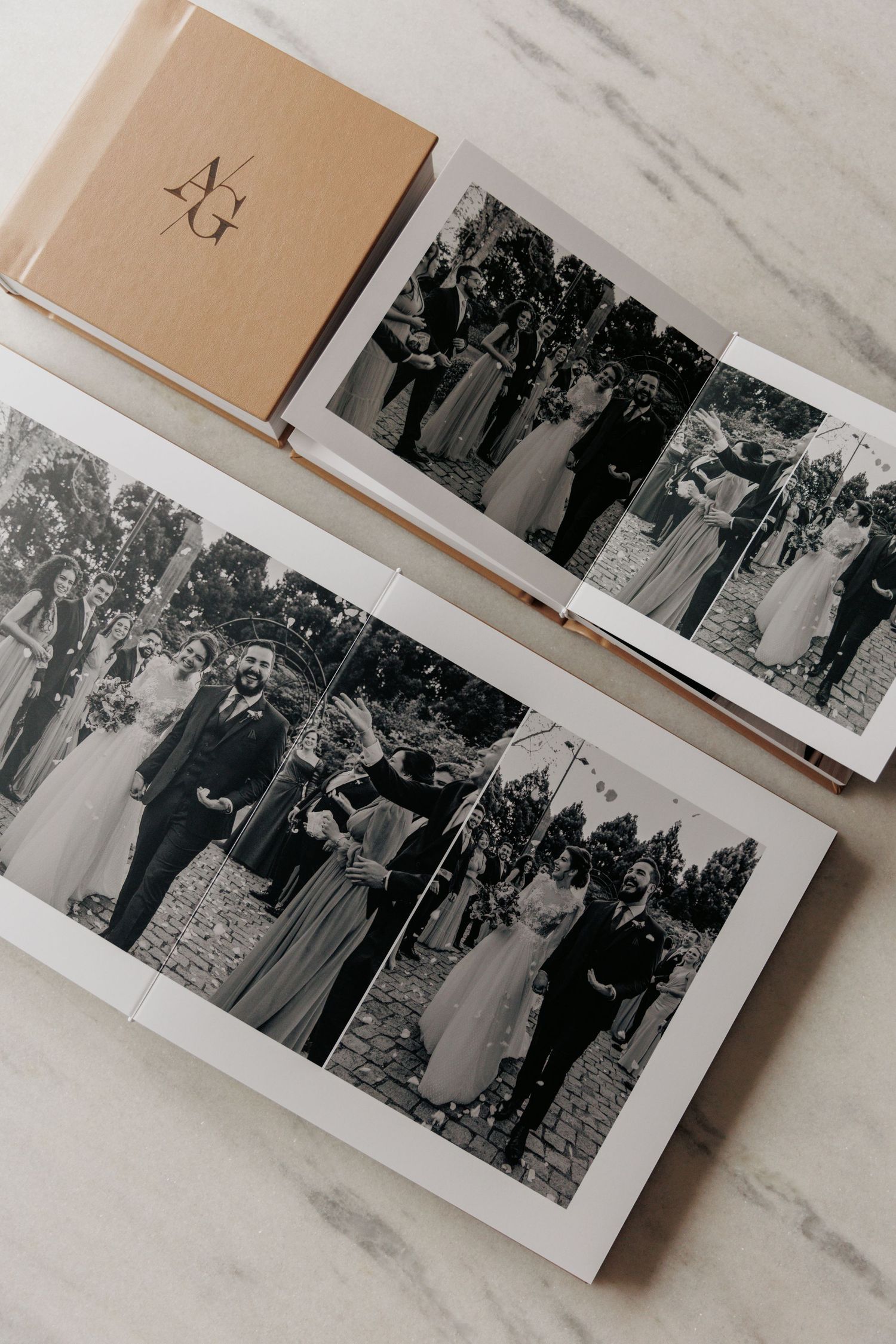 Open wedding photo album displaying black and white ceremony photos beside a monogrammed tan linen cover book on marble.