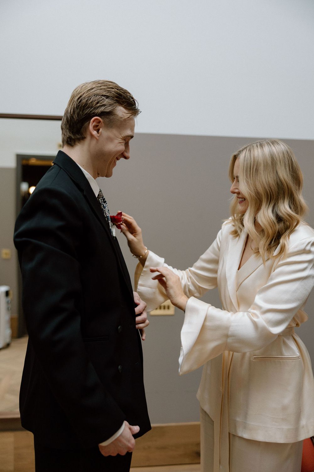 Bride in white suit pins boutonniere on groom in black tuxedo before wedding ceremony.
