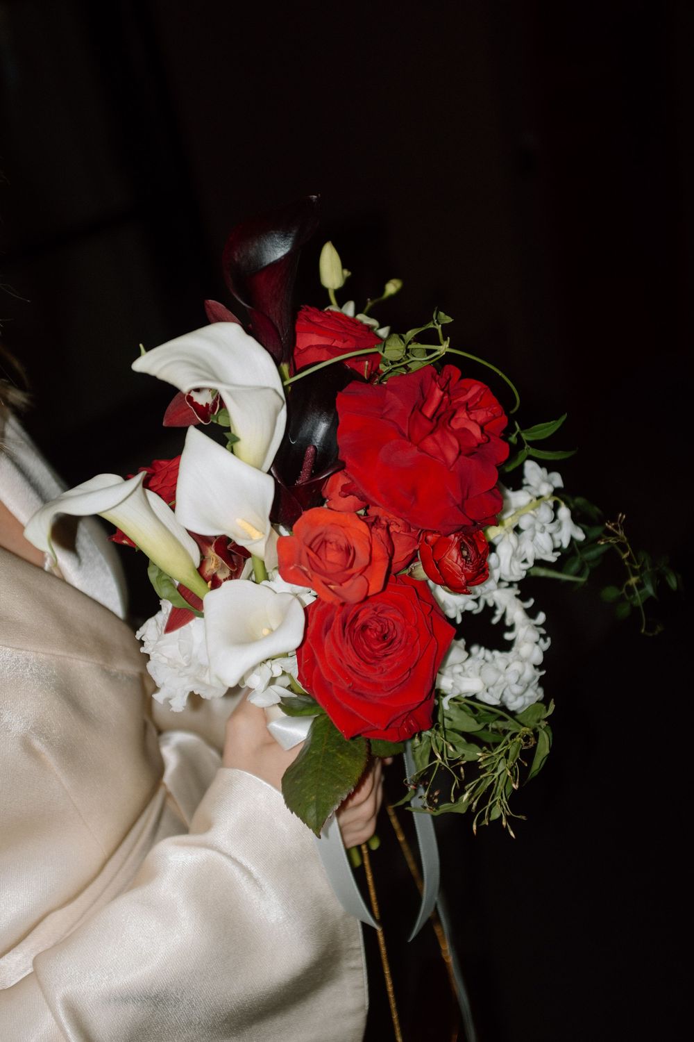 Bride holding elegant wedding bouquet with red roses and white calla lilies in satin dress.