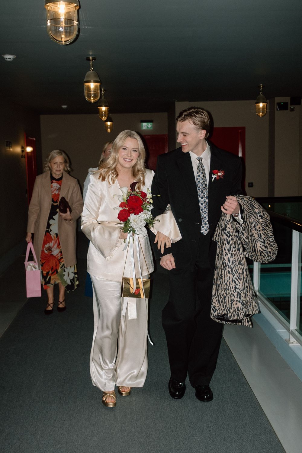 Smiling bride in satin pantsuit walking with groom in black suit at wedding venue.