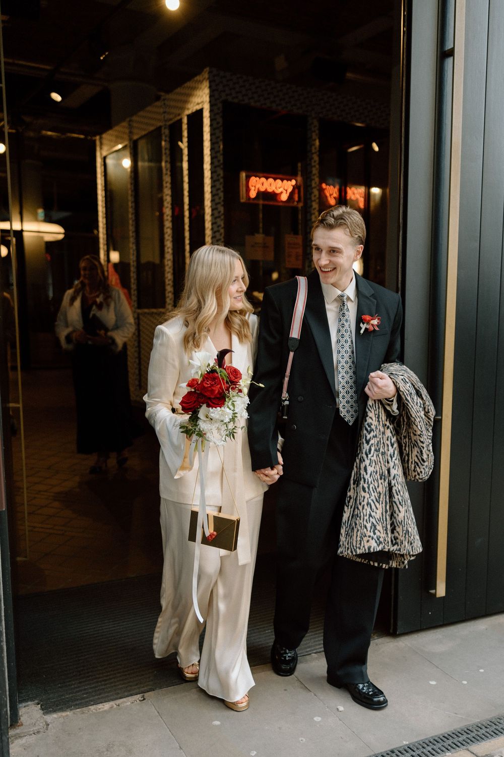 A smiling bride in a white pantsuit holding red roses walks with a groom in a black tuxedo outside a venue.