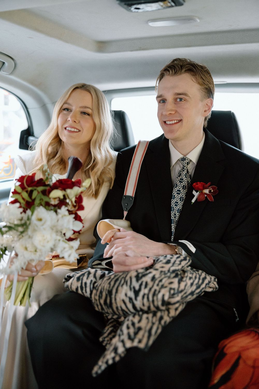 Bride and groom sitting in a car, smiling, with red and white wedding bouquet and black suit with floral boutonniere.