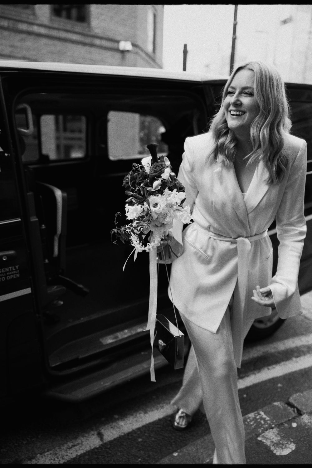 Smiling bride in white blazer suit holding bouquet stepping out of black taxi cab on wedding day.