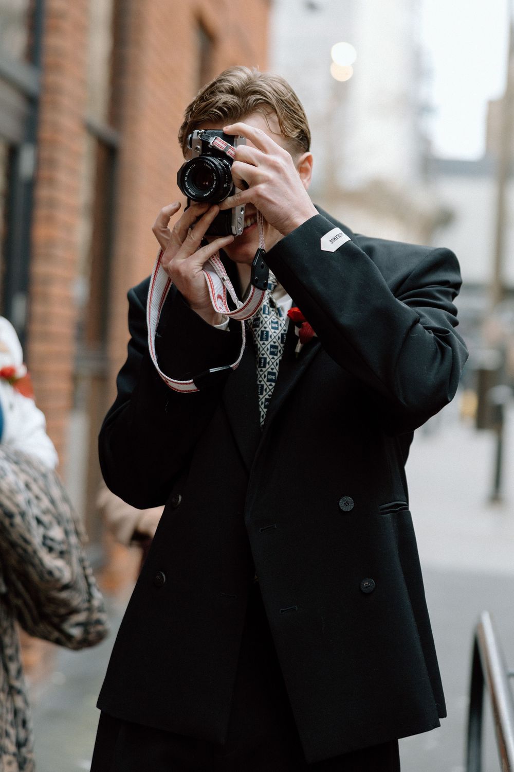 A stylish young man in a black blazer photographs with a film camera on a city street.