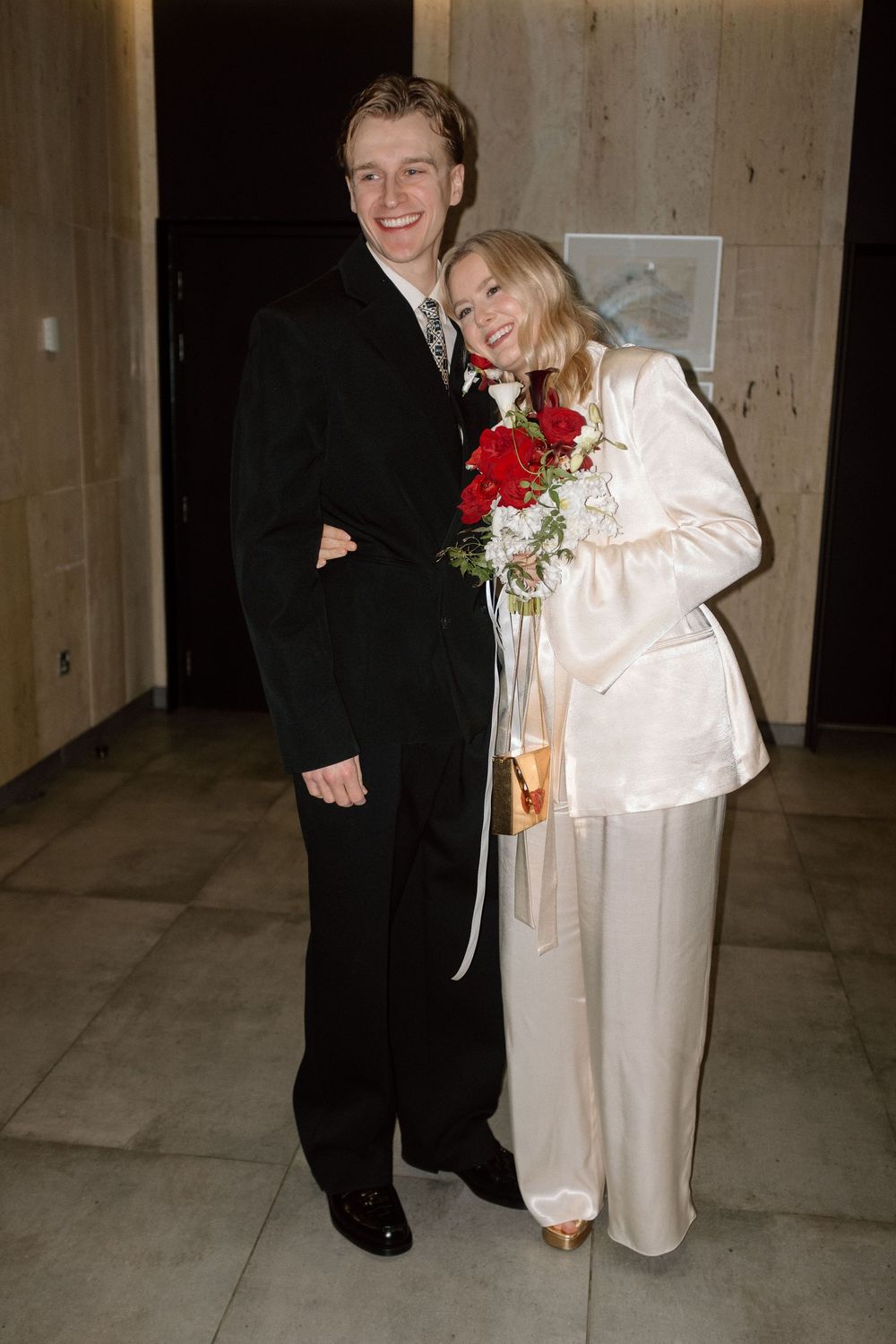 Newlyweds posing in a hallway, groom in black suit and bride in white pantsuit holding red roses bouquet.