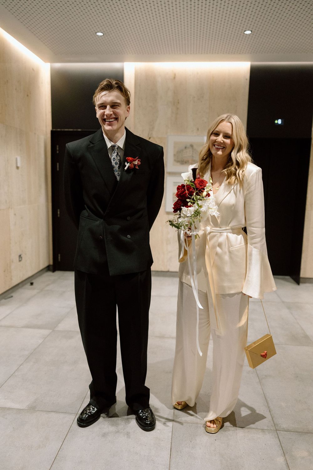 Smiling newlyweds pose indoors, groom in black suit with boutonniere, bride in white pantsuit holding red roses bouquet.
