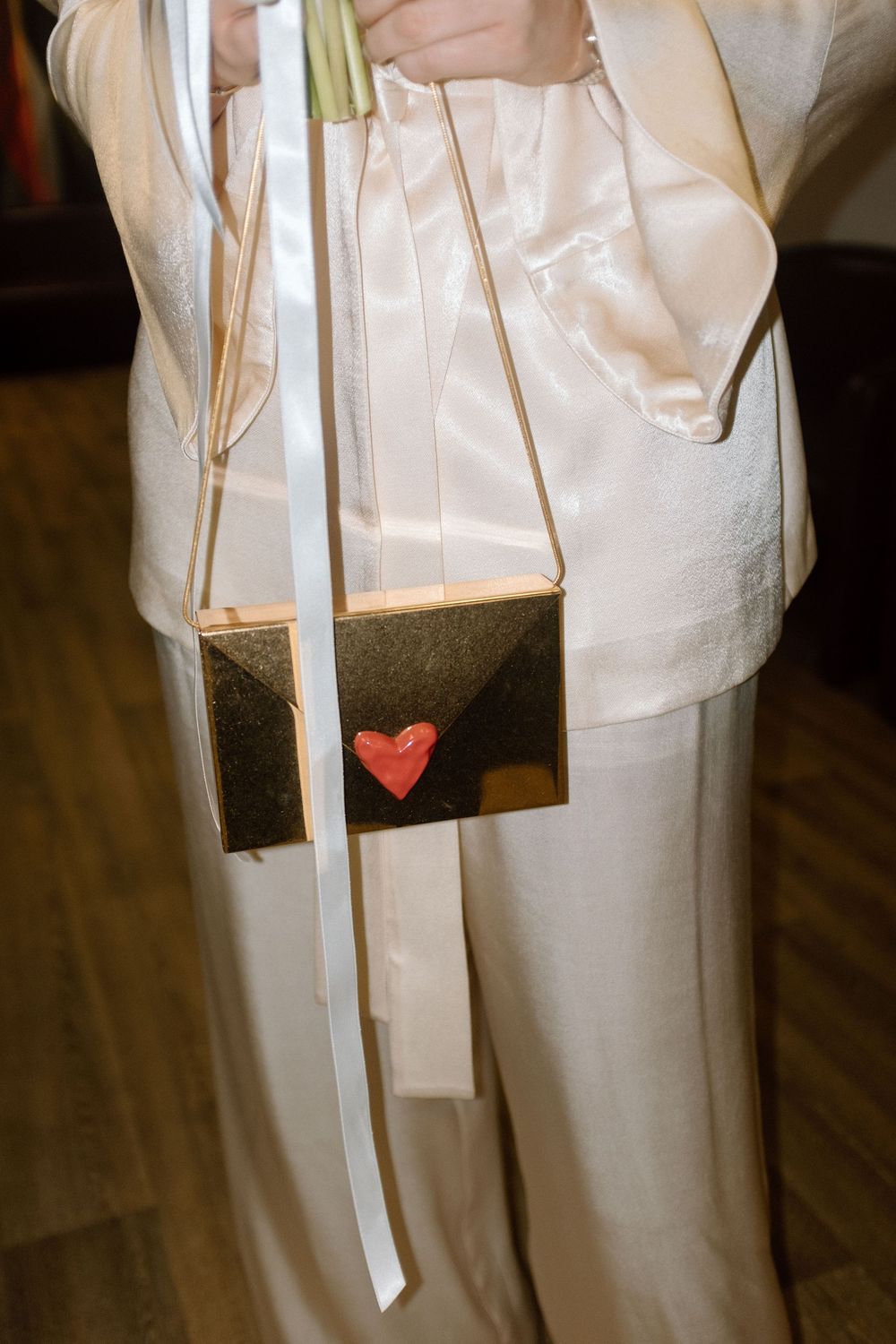 Close-up of bride in white satin pantsuit holding gold clutch purse decorated with red heart detail.