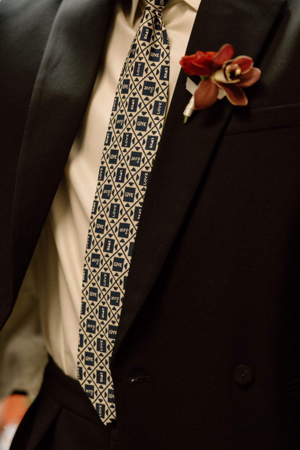 Close-up of a groom wearing a black suit with a patterned tie and red floral boutonniere on his lapel.