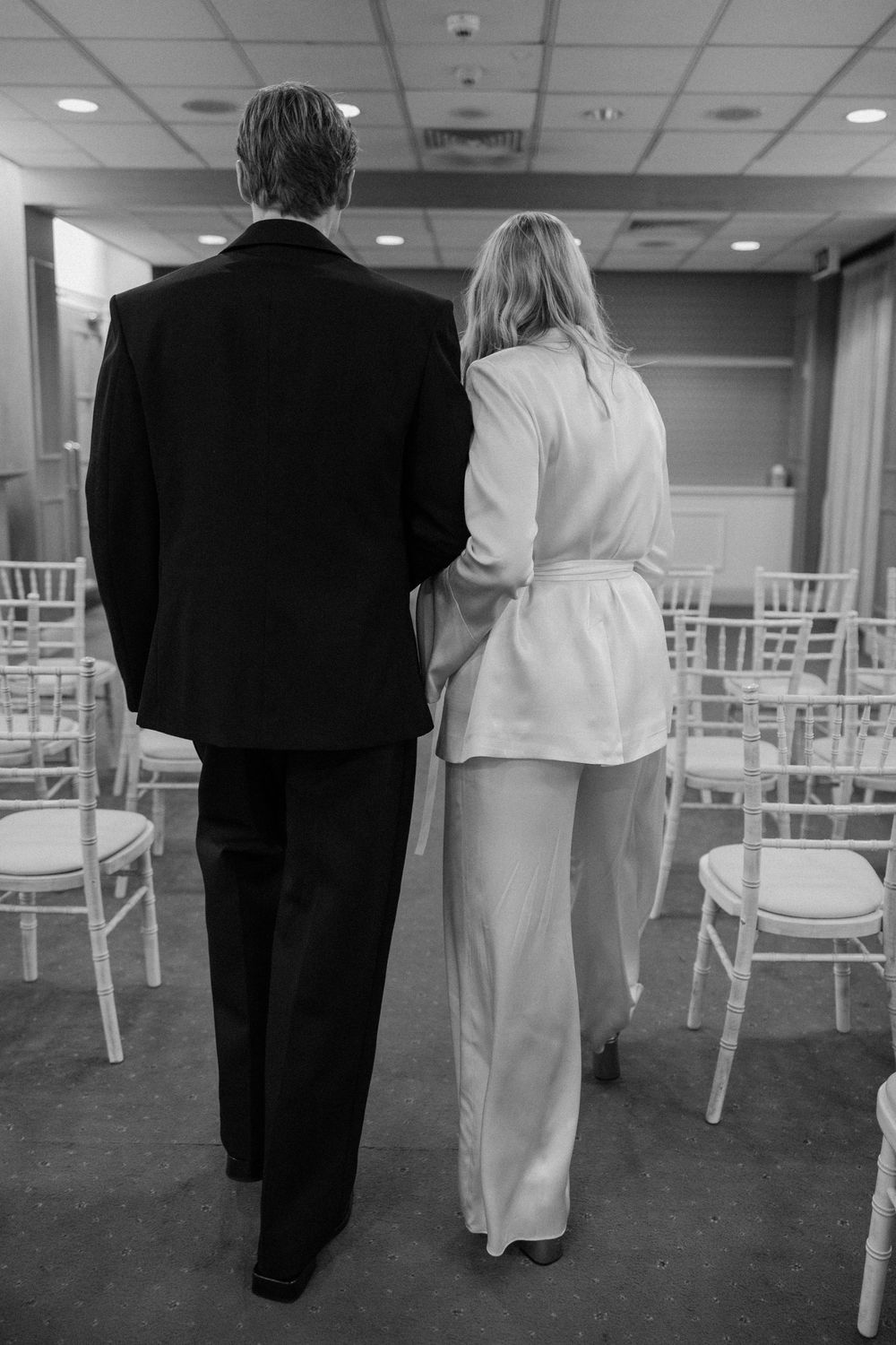 Black and white photo of bride and groom walking arm in arm through empty wedding ceremony venue with white chairs.