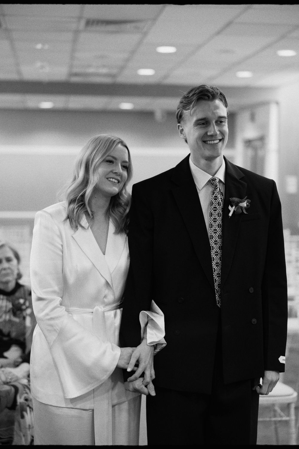 Smiling bride in white suit and groom in black suit holding hands during wedding ceremony.