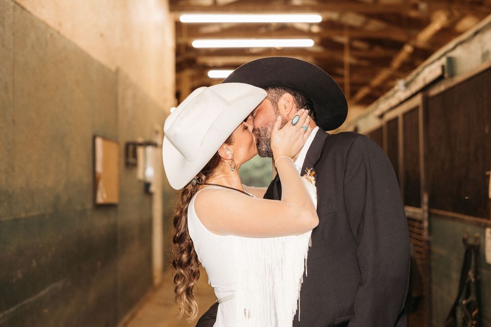 Couple sharing a kiss in a rustic barn, bride in white fringe dress and cowboy hat, groom in black hat and jacket.