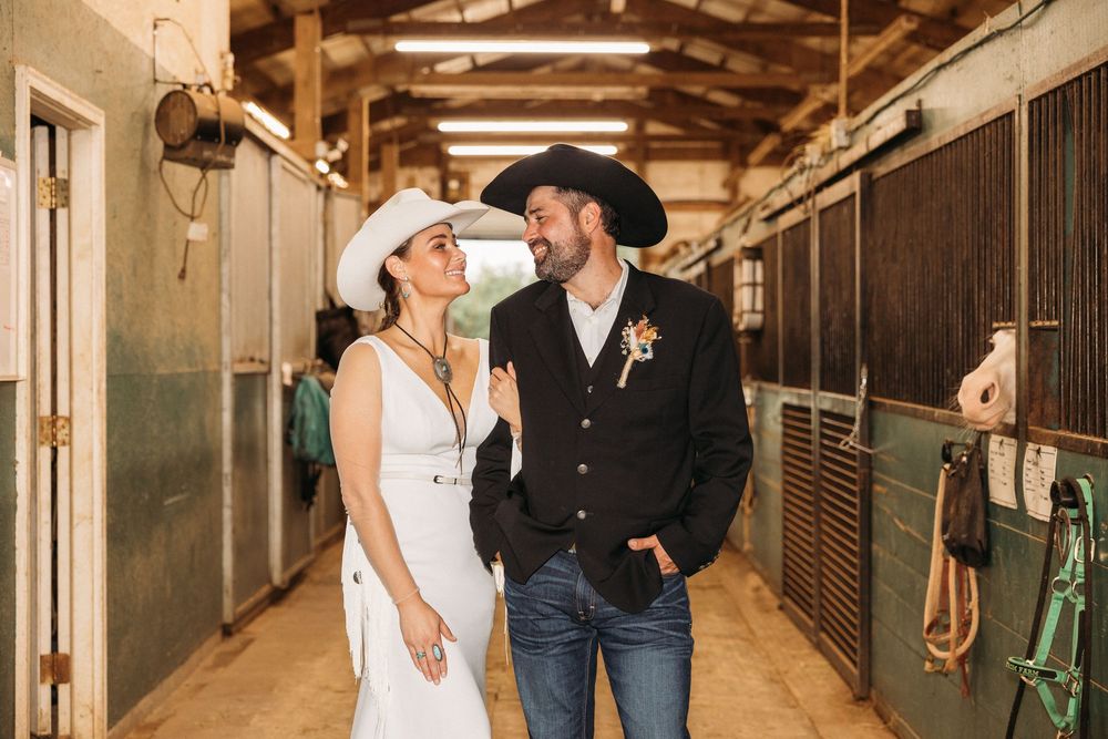 Couple in cowboy hats smiling at each other in a rustic barn aisle, wearing western wedding attire.