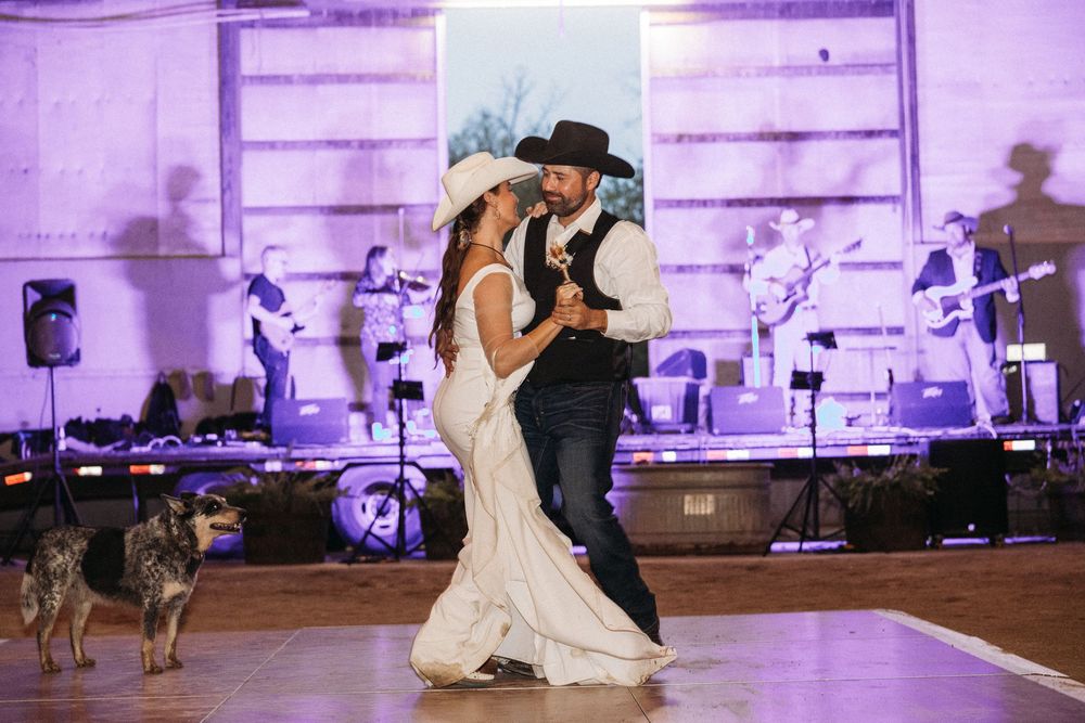 Bride and groom share first dance at rustic wedding reception with live band performing on stage and dogs nearby.