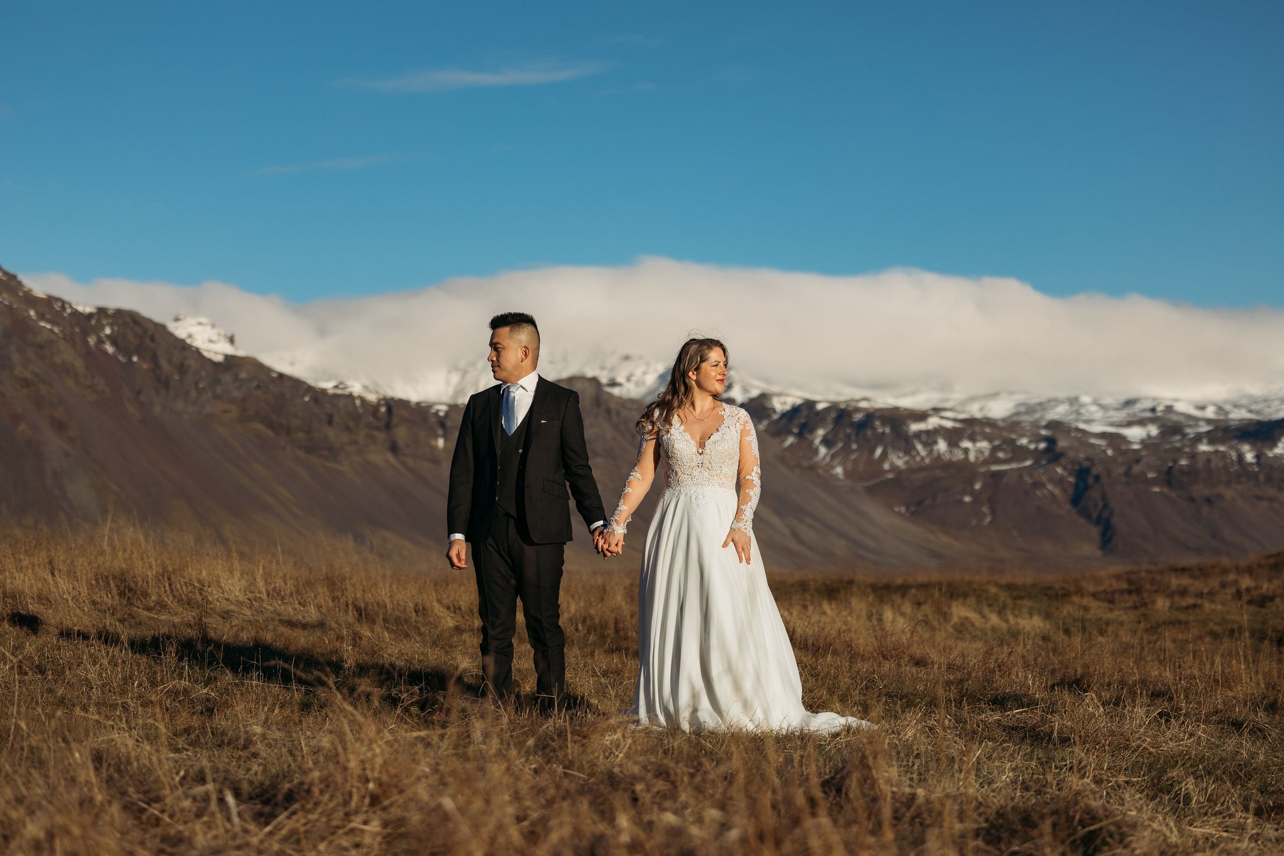 Couple in wedding attire walk hand in hand through golden meadow with stunning snow-capped mountain range behind them.