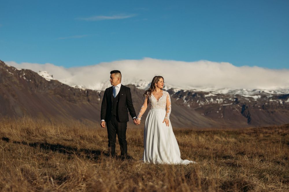 Couple in wedding attire walk hand in hand through golden meadow with stunning snow-capped mountain range behind them.