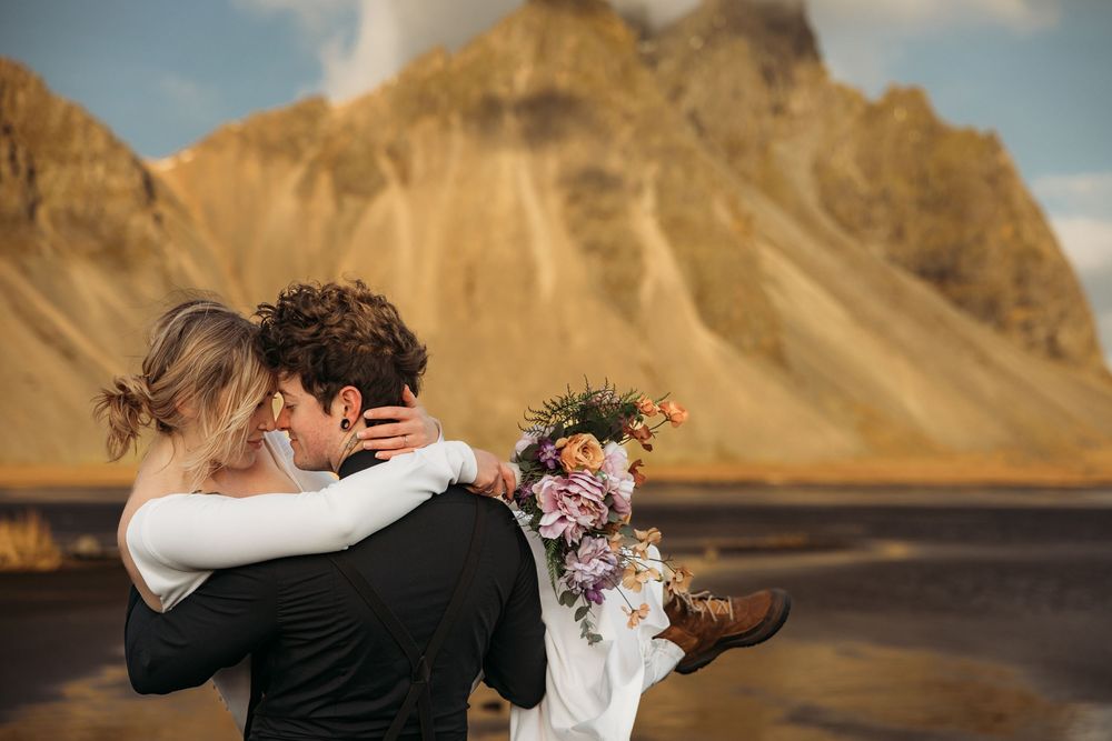 Couple embracing at Iceland's Vestrahorn mountain, groom lifting bride holding colorful floral bouquet on black sand beach.