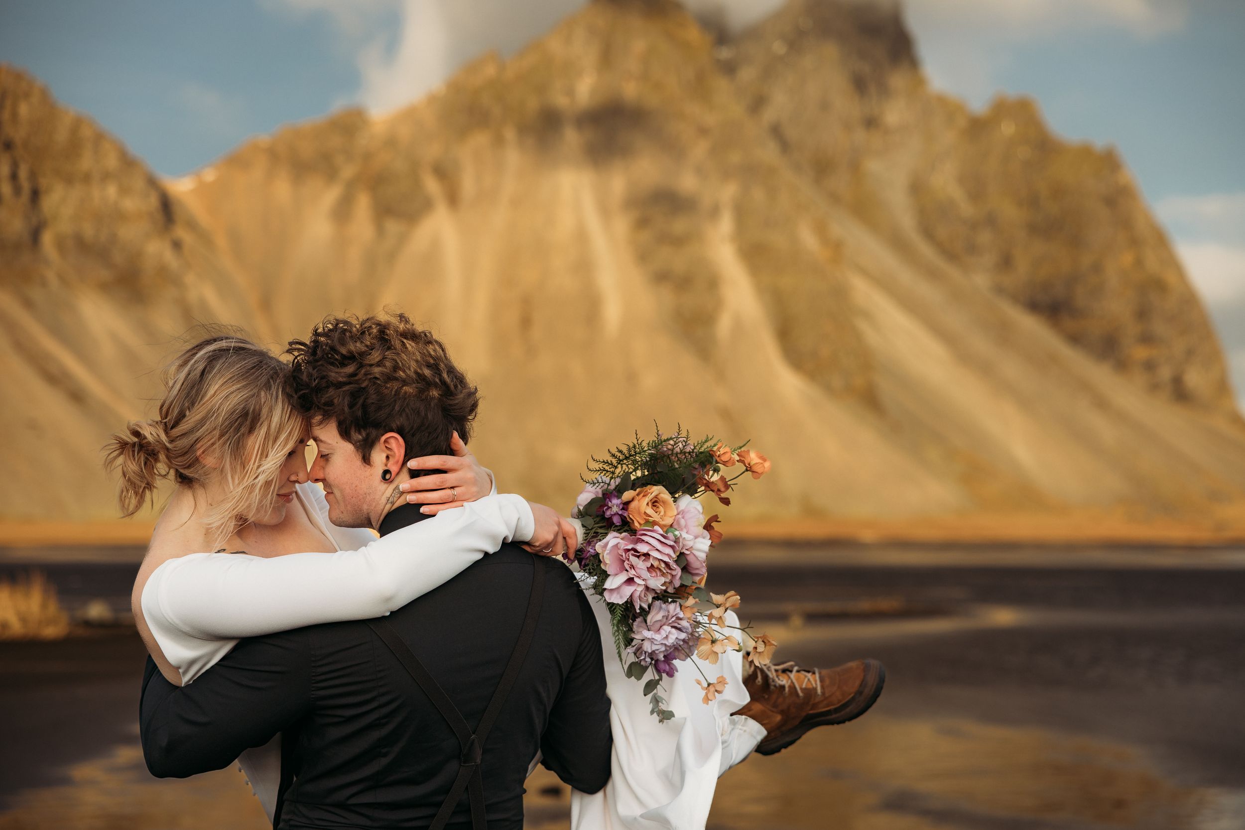 Couple embracing at Iceland's Vestrahorn mountain, groom lifting bride holding colorful floral bouquet on black sand beach.