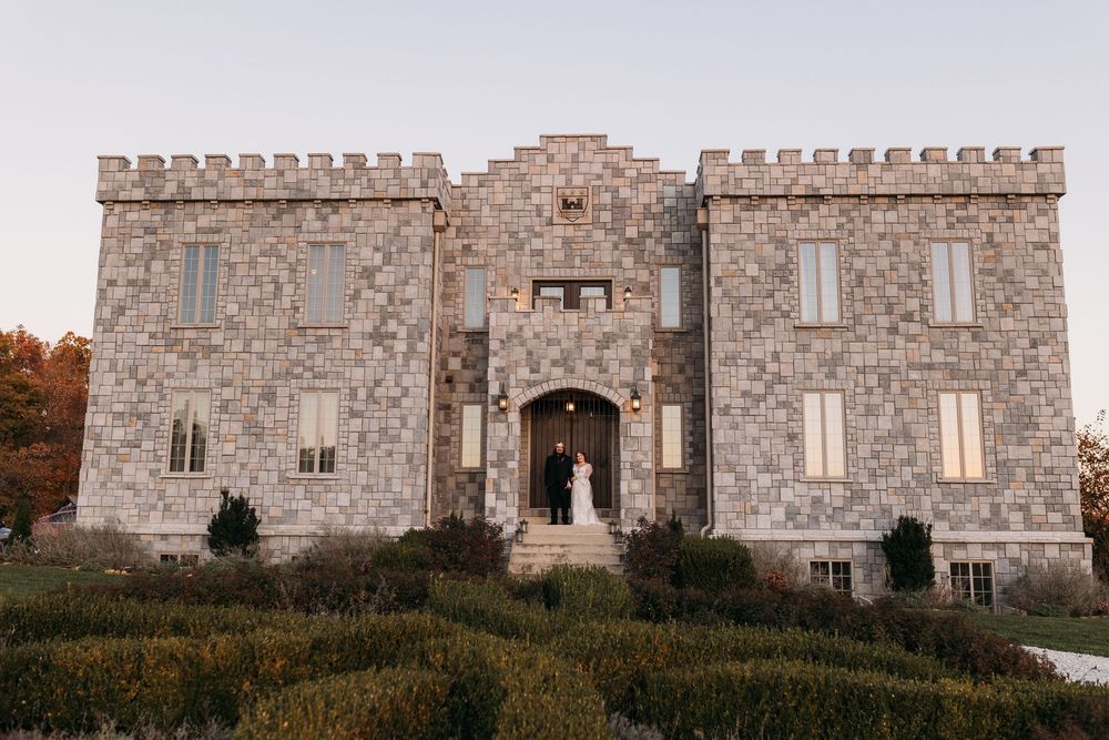 Couple poses in arched doorway of stone castle wedding venue with battlements and manicured hedges at sunset.