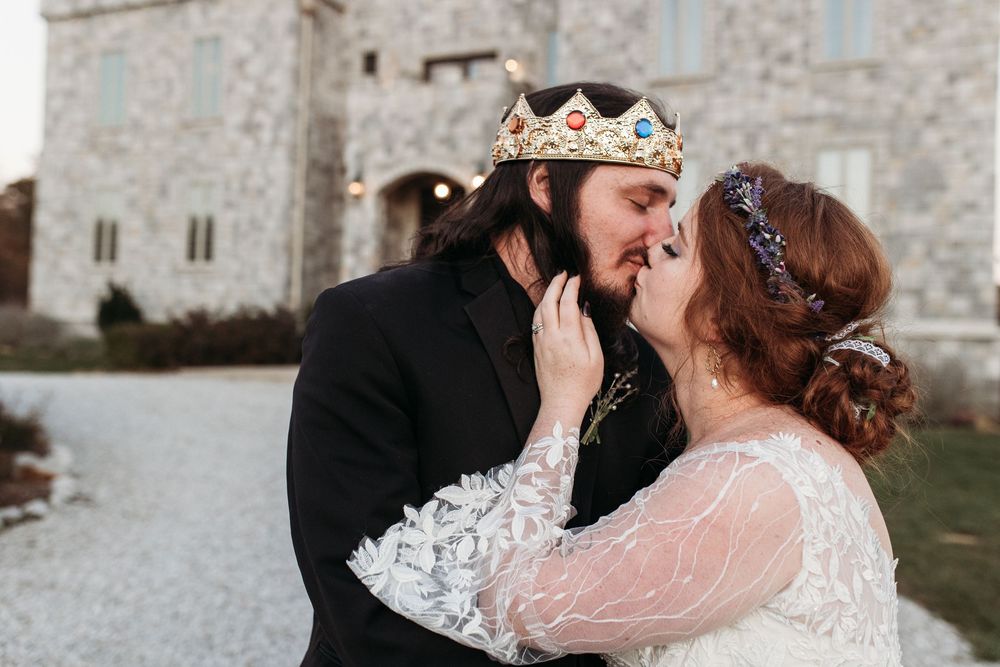 A couple in wedding attire kisses outside a castle, the groom wearing a jeweled crown and bride in a lace gown.