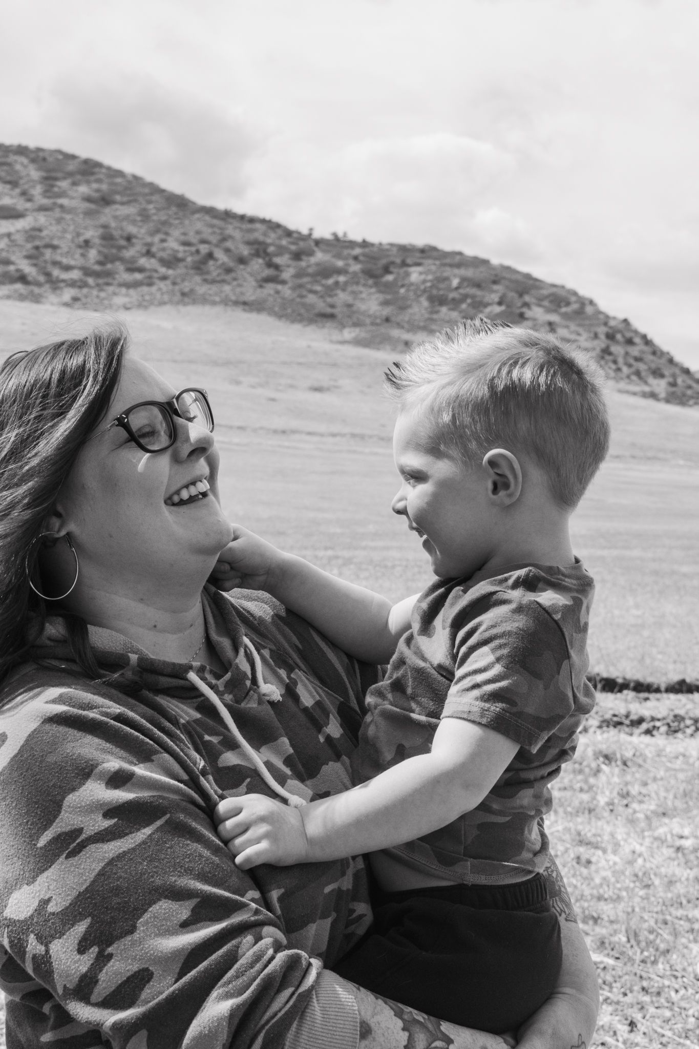 Red-haired woman hugging smiling young boy in camo shirt outdoors with mountain landscape background.