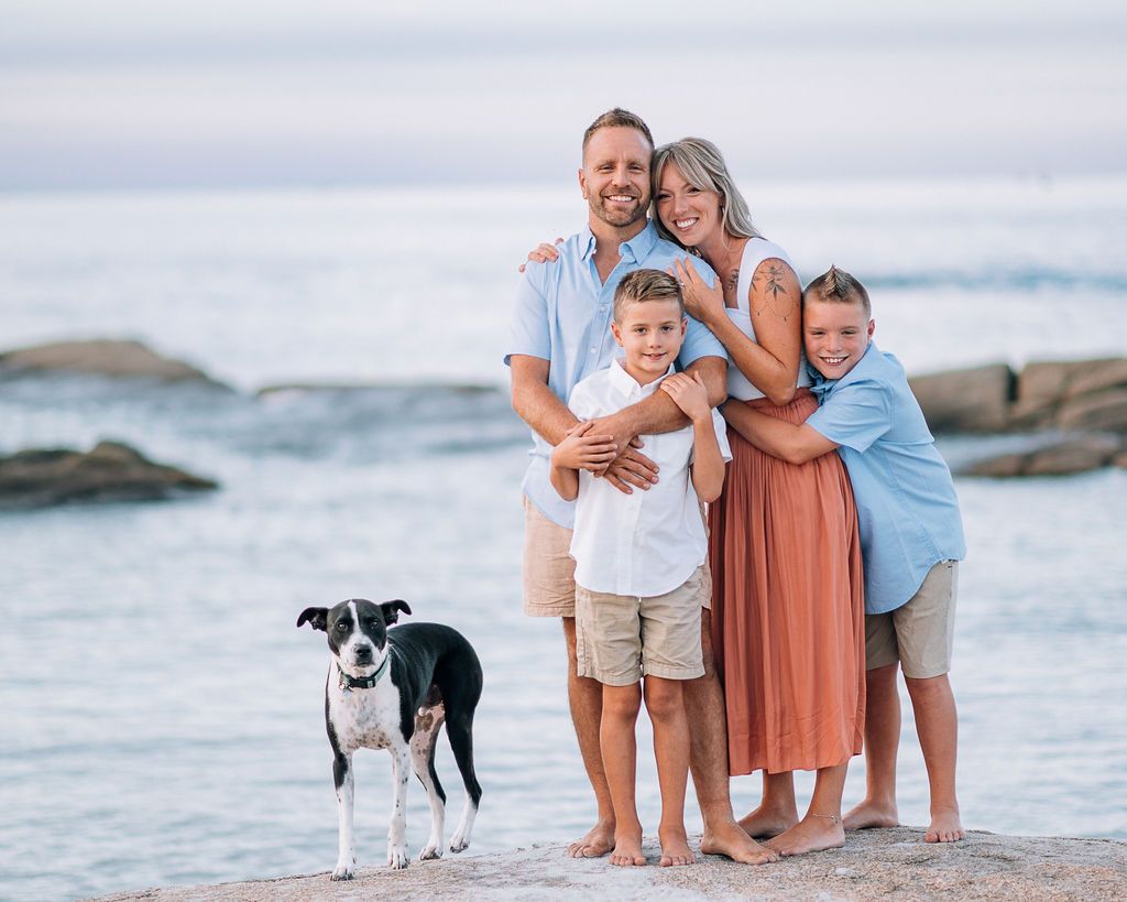 Family of four with a black and white dog posing on a sandy beach with ocean rocks in the background.