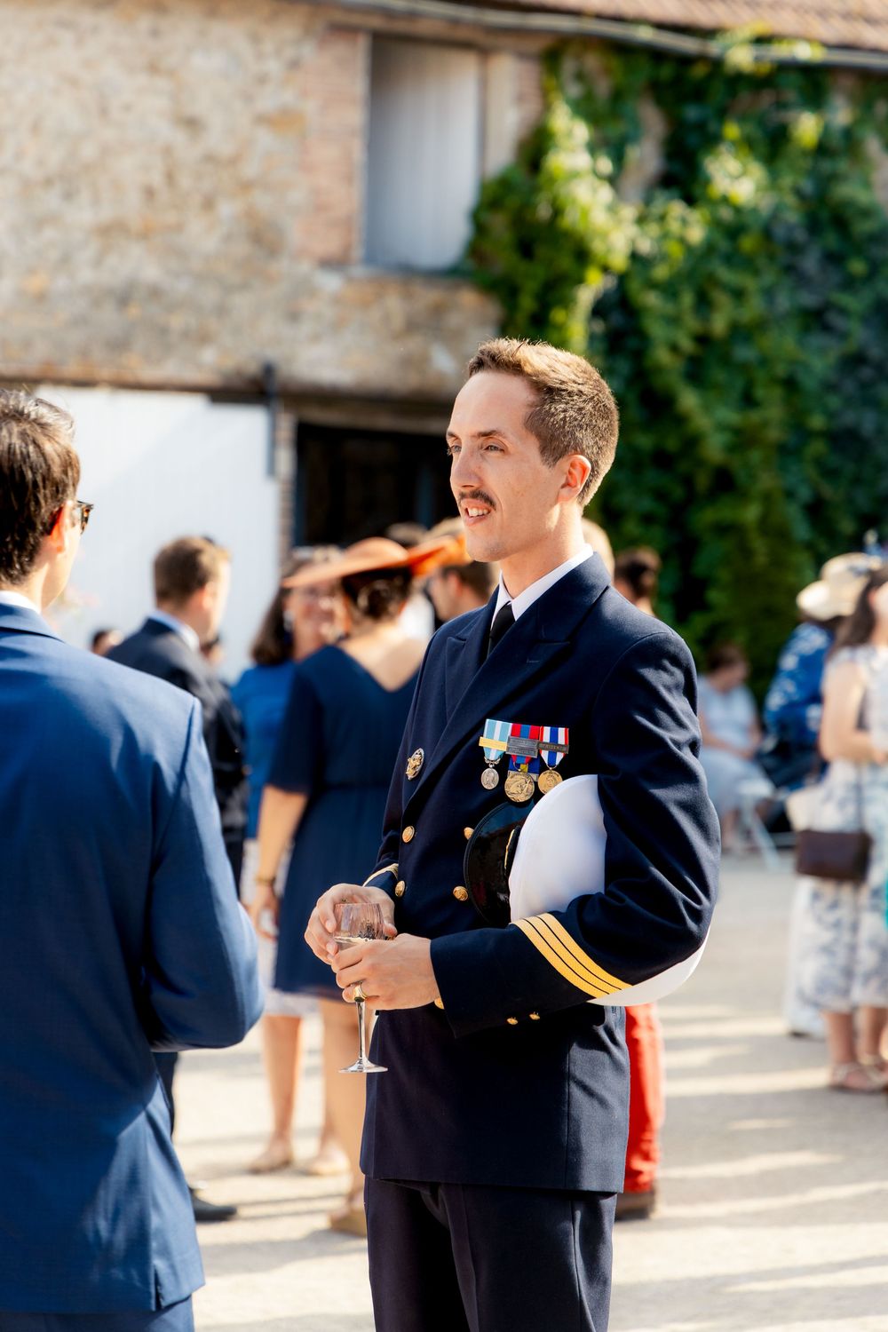 Military officer in dress uniform with medals holds champagne glass at outdoor social gathering.