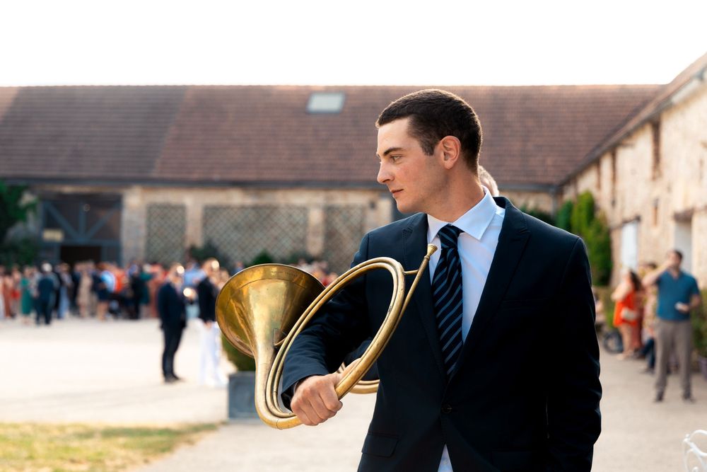 Young man in dark suit carrying a brass tuba outdoors at a countryside gathering.