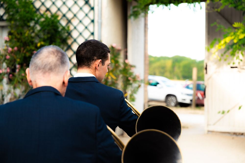 Two men in dark suits holding brass instruments wait near an outdoor venue entrance.