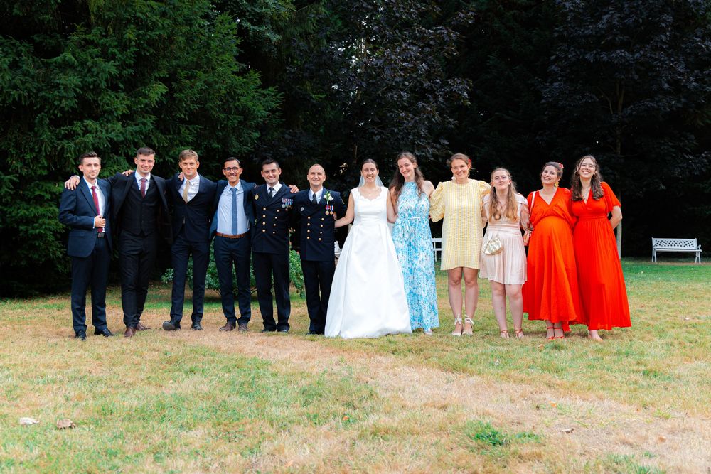 Wedding party poses outdoors on grass with trees, bride in white gown flanked by guests in formal attire and colorful dresses.