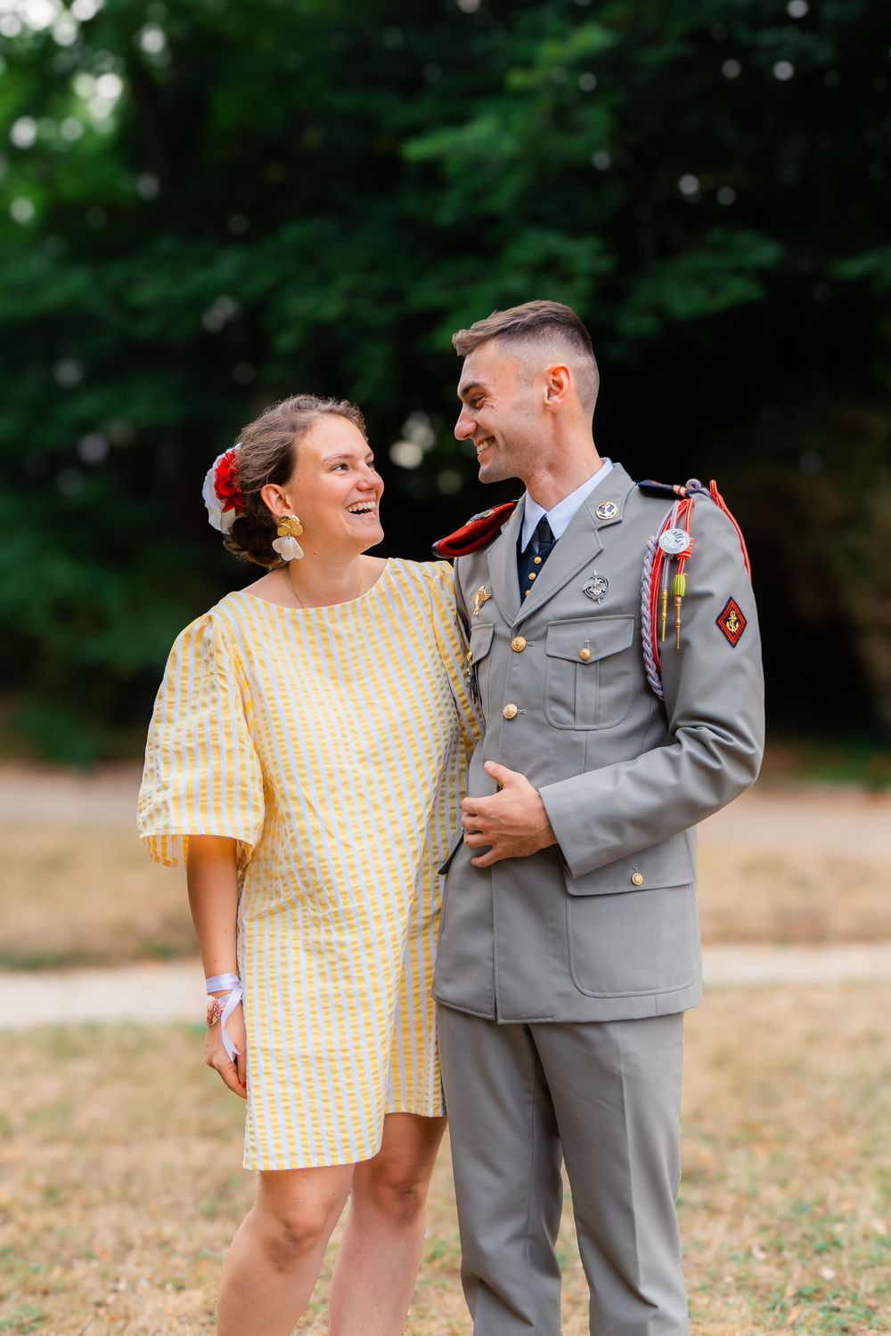 A smiling couple outdoors, woman in yellow dress and man in military uniform with medals, sharing a joyful moment.