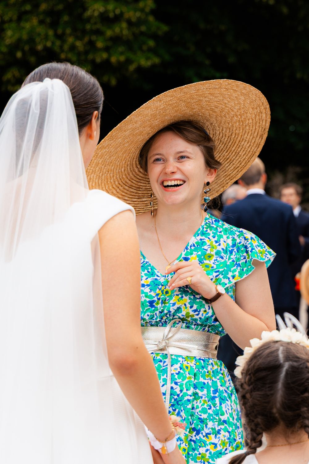 A laughing woman in a straw hat and floral dress chats with a bride in a white veil at an outdoor wedding.