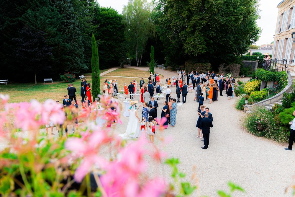 Aerial view of wedding guests gathering in elegant garden with pink flowers, bride in white gown, and lush greenery.