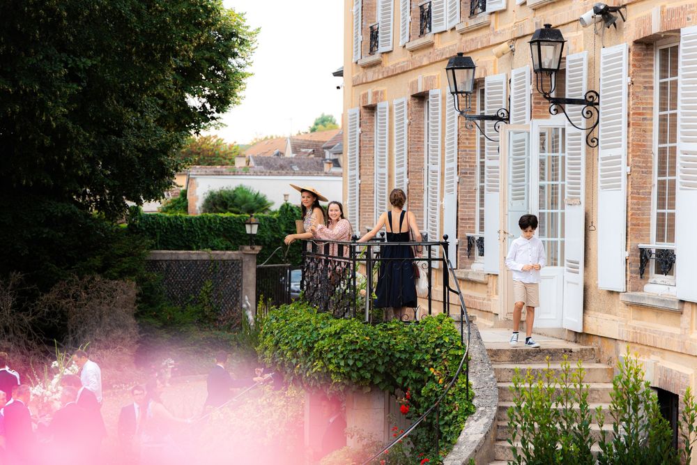 Elegant French chateau with guests on a balcony surrounded by lush greenery and classic shuttered windows.