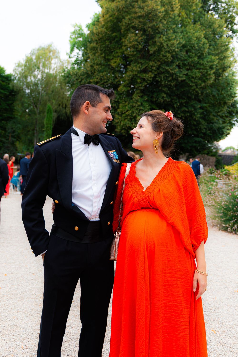 Pregnant woman in red gown with man in military tuxedo smiling at each other outdoors.
