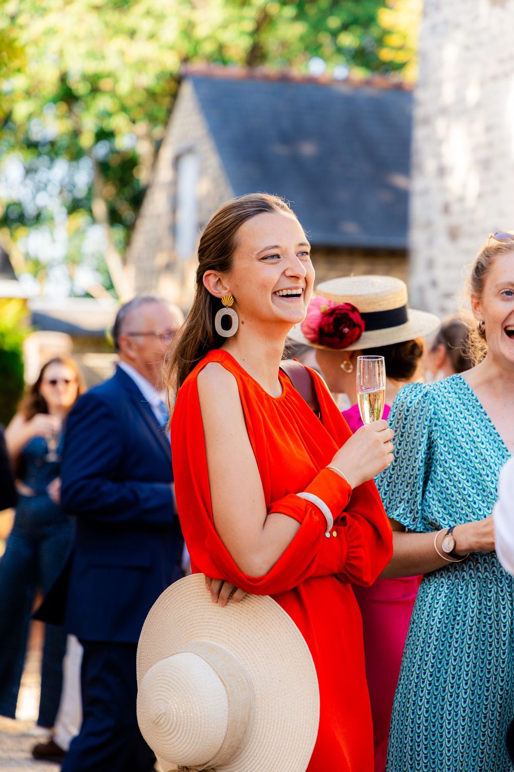 A laughing woman in a red dress holds a champagne glass and sun hat at an outdoor garden party.