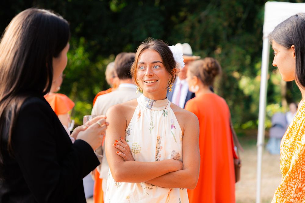 Woman in white halter dress smiling with arms crossed at an outdoor social gathering with guests in orange attire.