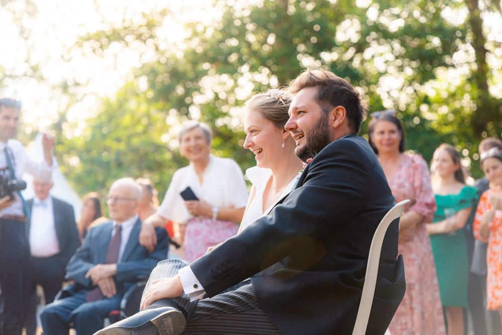 A joyful couple sits together laughing at an outdoor wedding ceremony surrounded by smiling guests in a sunlit garden.