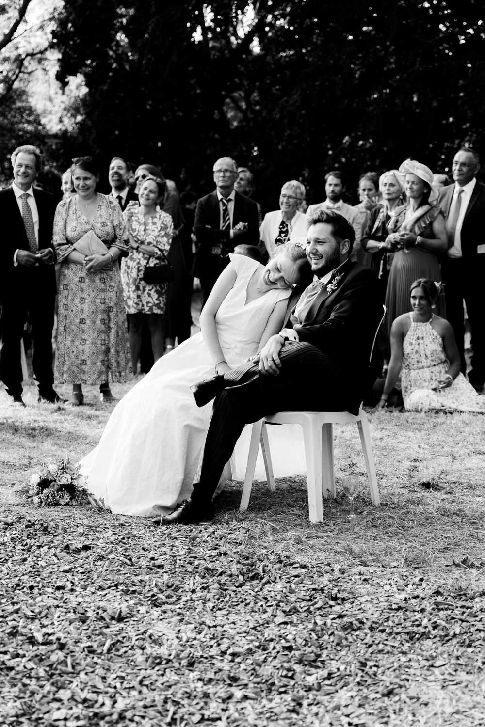 Black and white wedding photo of bride leaning on groom seated outdoors, guests watching ceremony behind them.