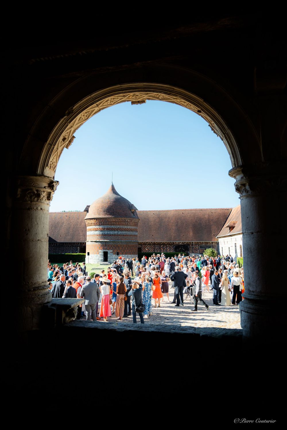 Elegant outdoor wedding reception in a historic French chateau courtyard, viewed through a stone archway on a sunny day.