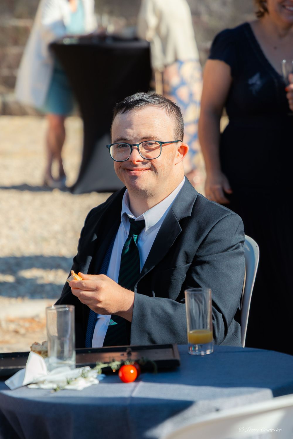Smiling man with Down syndrome in suit and tie seated at outdoor event table with drinks.