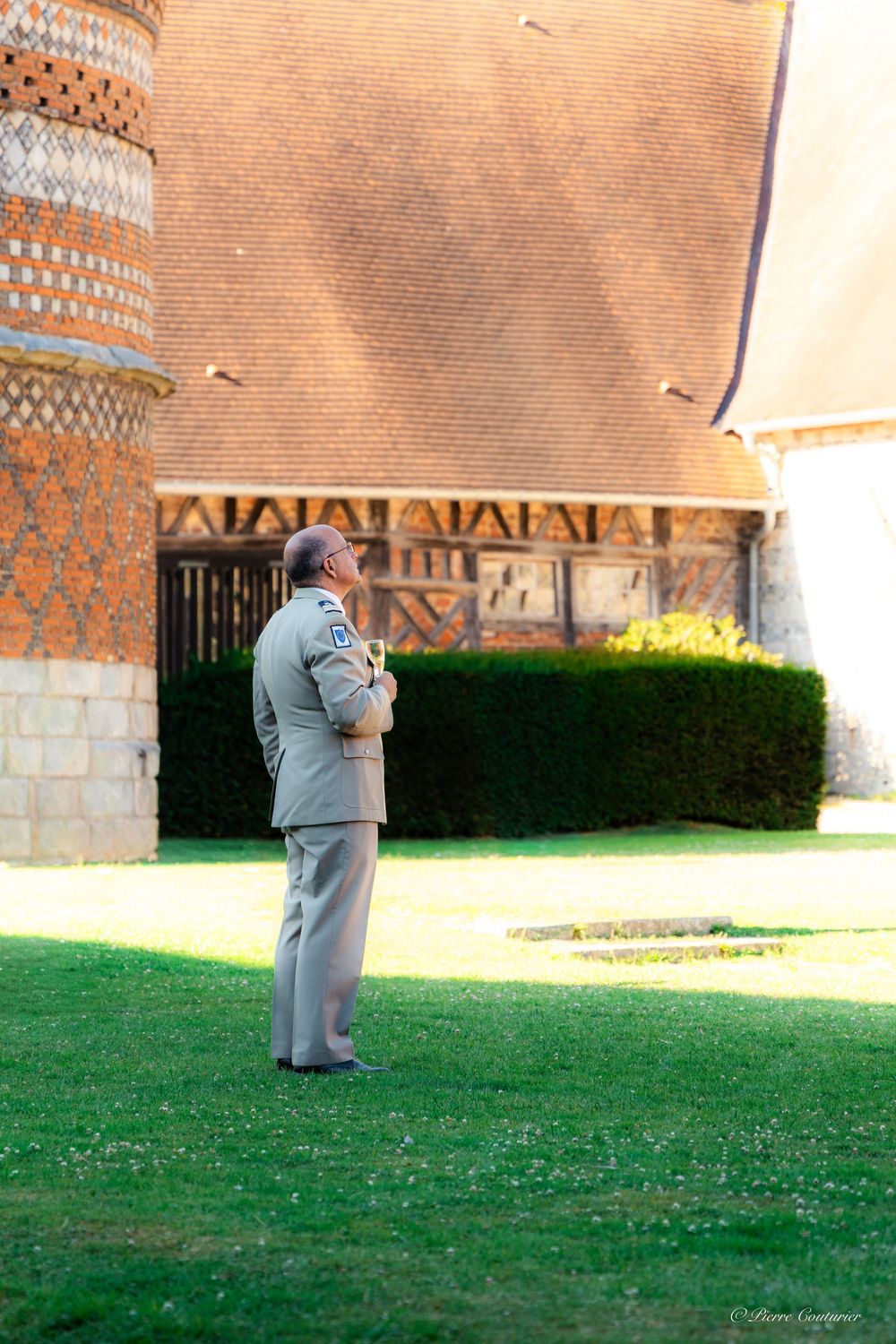 Military officer in uniform stands on green lawn before historic brick building with decorative roof patterns.
