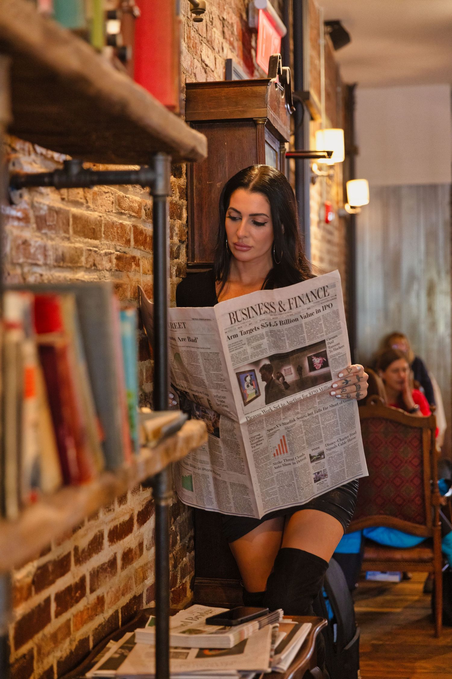 Woman reading Business & Finance newspaper in a cozy cafe with brick walls and bookshelves.