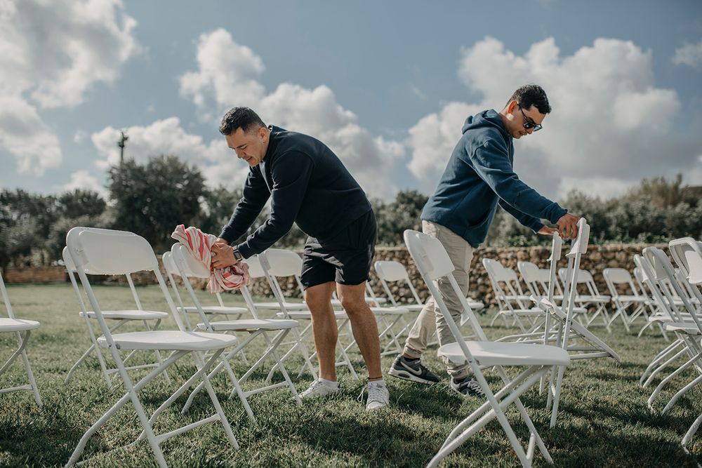 Two men arranging white folding chairs on grass outdoors for a wedding ceremony under a cloudy sky.