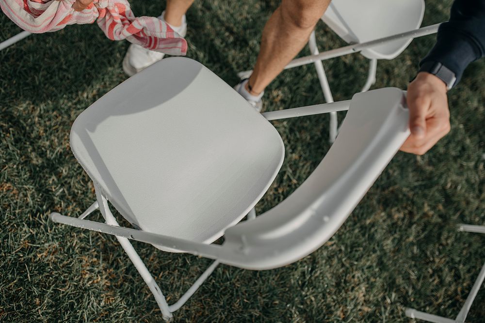 Two people setting up white folding chairs on grass at an outdoor event or wedding ceremony.