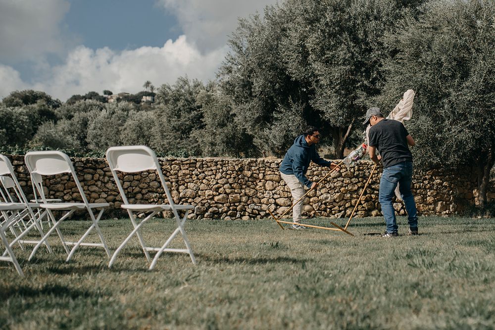 Two people setting up white folding chairs on a green lawn near a stone wall with olive trees in the background.