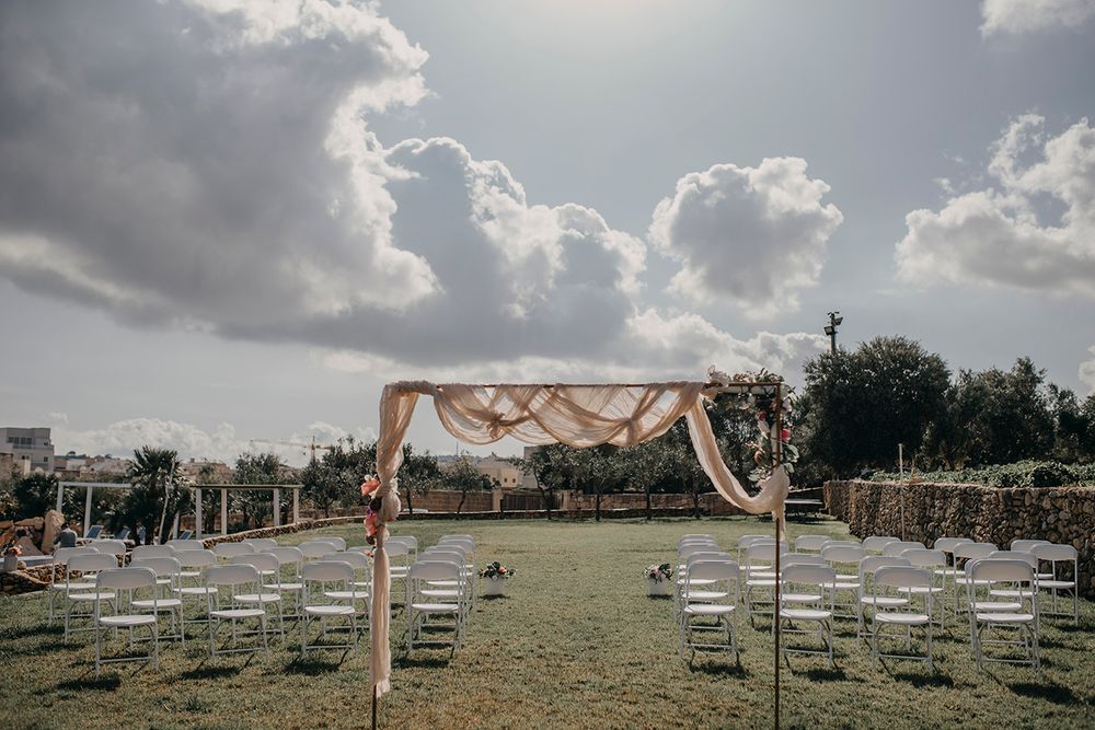 Outdoor wedding ceremony setup with white chairs, floral arch, and draped fabric under a cloudy sky.